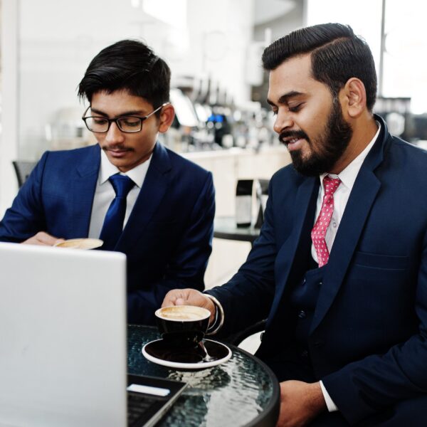 two-indian-business-man-suits-sitting-office-cafe-looking-laptop-drinking-coffee-min